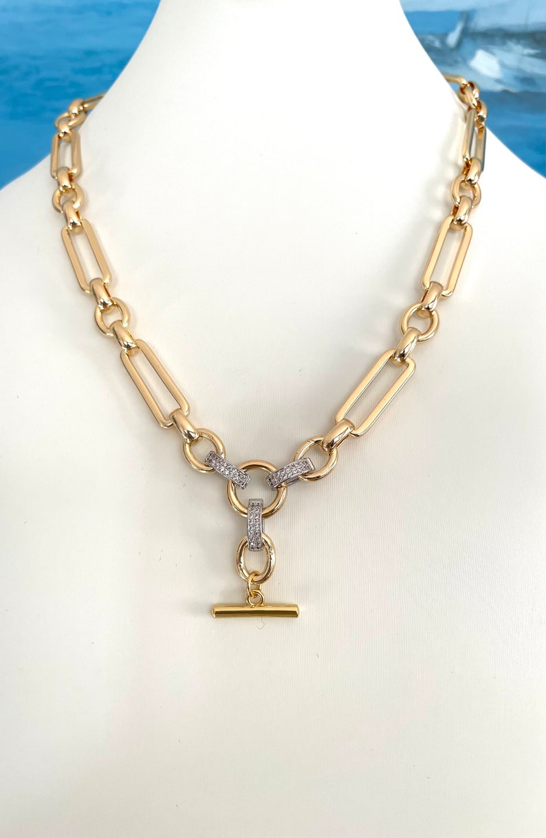 May include: A gold chain necklace with a toggle clasp. The chain is made of thick, rectangular links. The toggle clasp is decorated with small, clear stones.