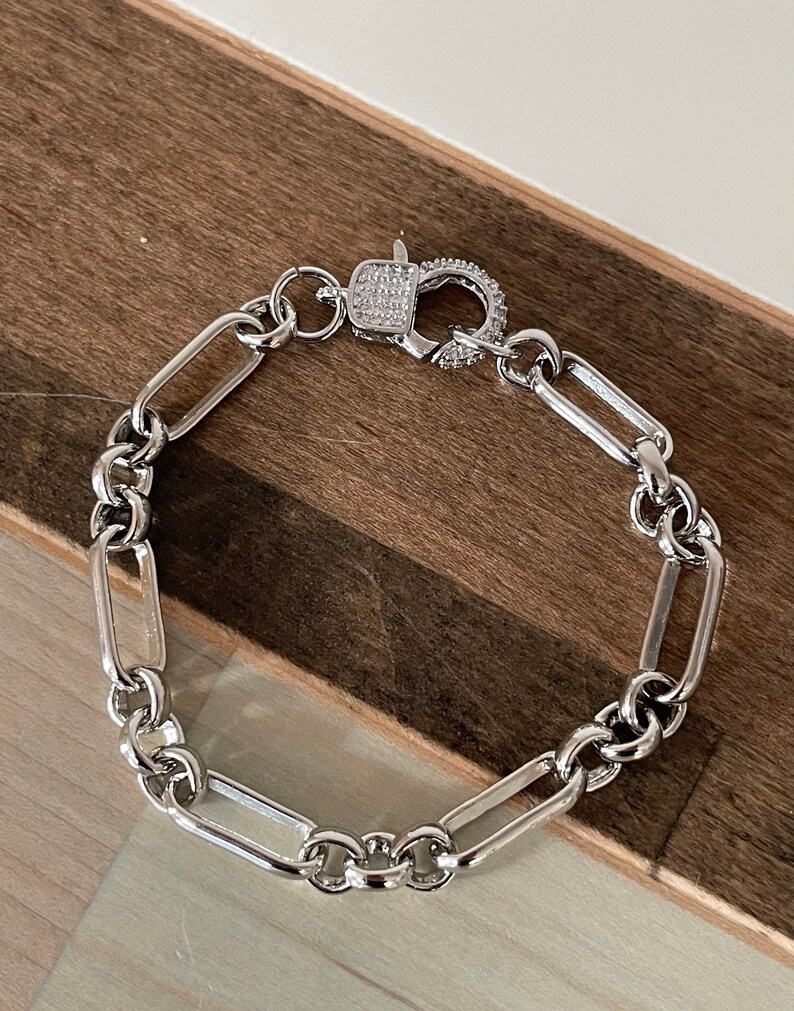 May include: A silver-toned bracelet with a rectangular link chain design. The clasp is encrusted with small, clear stones. The bracelet is displayed on a wooden surface.