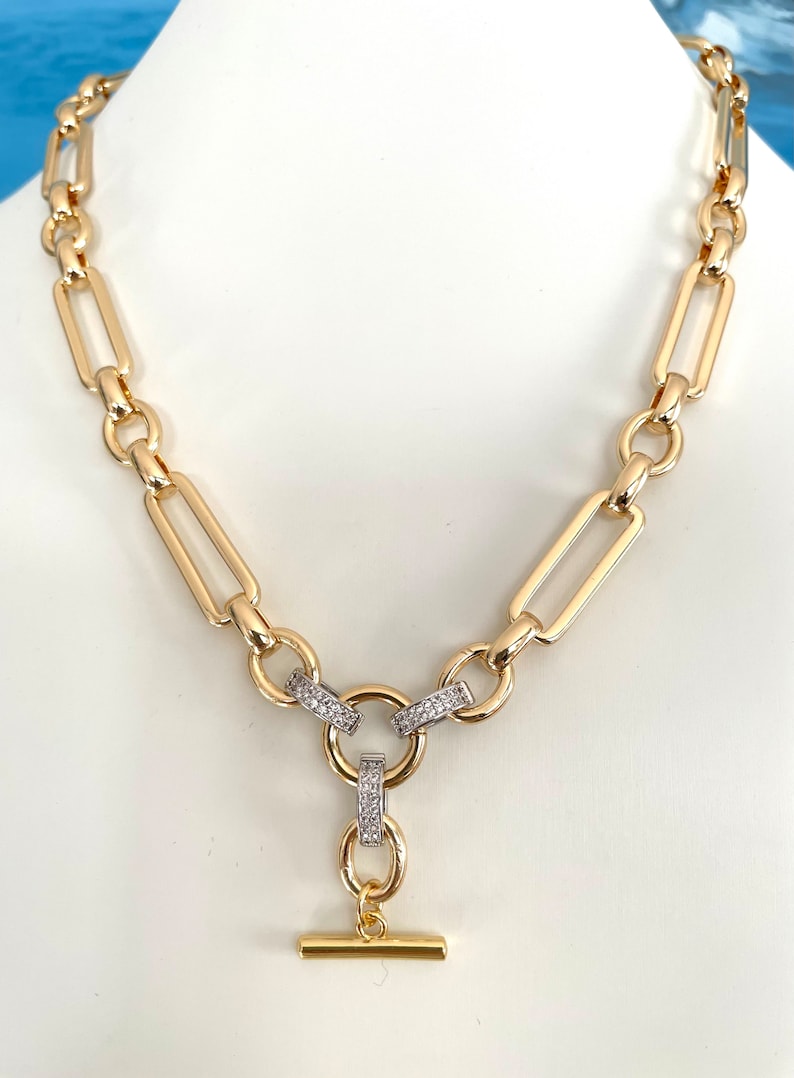 May include: A gold chain necklace with a toggle clasp. The chain is made of large, rectangular links. The toggle clasp is decorated with small, clear stones.