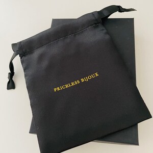 May include: A black satin drawstring bag with the text "PRICELESS BIJOUX" printed in gold lettering. The bag is sitting on top of a black box.