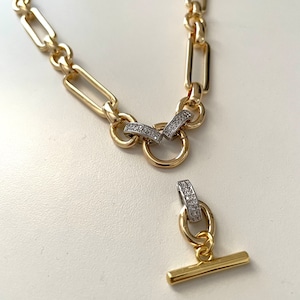 May include: A gold chain necklace with a toggle clasp. The chain is made of rectangular links and features small clear stones on the clasp.