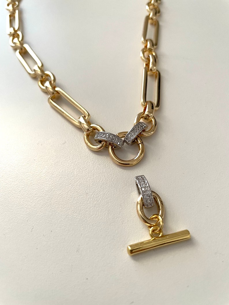 May include: A gold chain necklace with a toggle clasp. The chain is made of rectangular links and features small clear stones on the clasp.