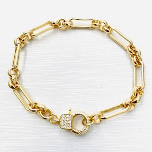 May include: Gold-toned bracelet featuring a mix of elongated and rounded links. The clasp is a square shape with a circle, both encrusted with small, clear stones. The bracelet is displayed on a white surface.