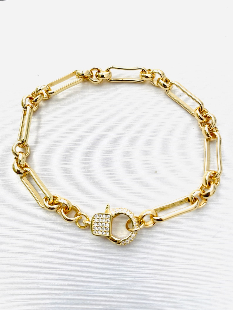 May include: Gold-toned bracelet featuring a mix of elongated and rounded links. The clasp is a square shape with a circle, both encrusted with small, clear stones. The bracelet is displayed on a white surface.