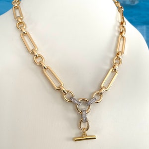 May include: A gold chain necklace with a large, chunky link design. The necklace has a T-bar clasp with a small, round charm that is encrusted with clear cubic zirconia stones.