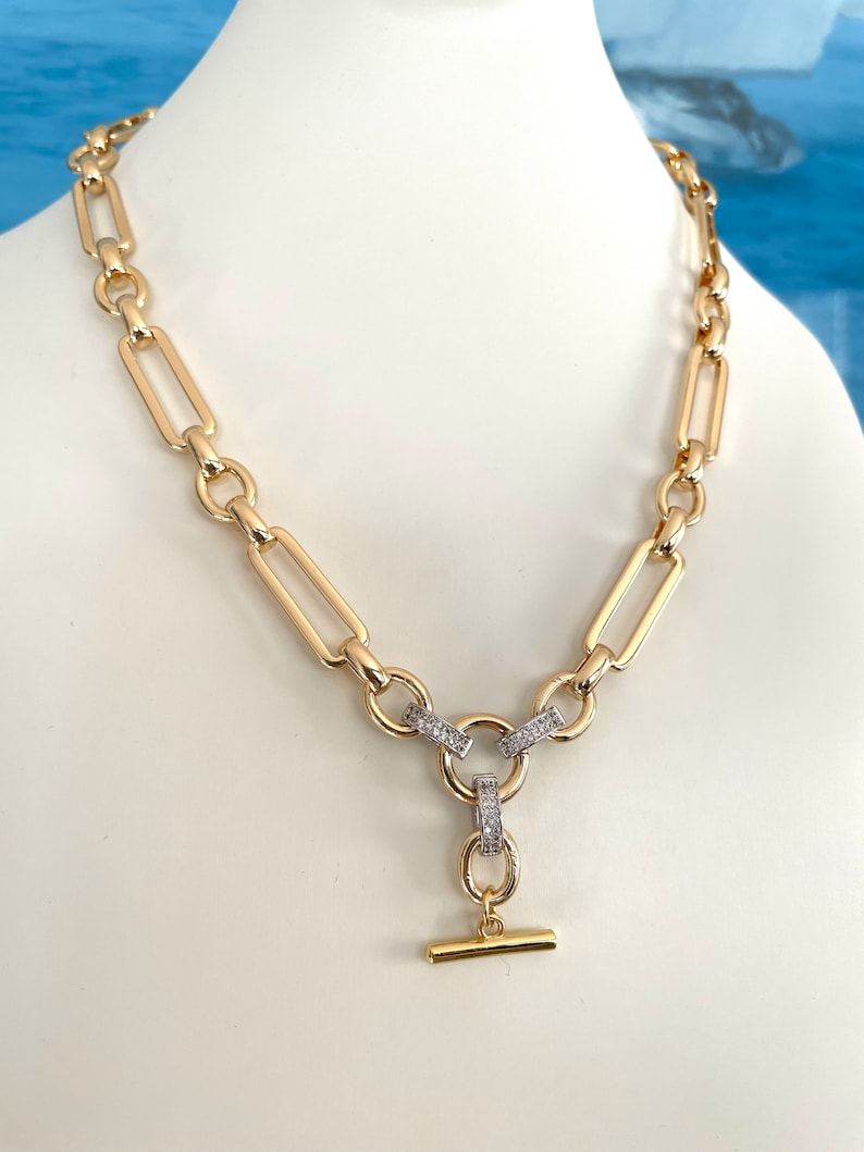 May include: A gold chain necklace with a large, chunky link design. The necklace has a T-bar clasp with a small, round charm that is encrusted with clear cubic zirconia stones.