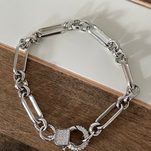 May include: Silver-toned chain bracelet with a rectangular link design. The bracelet features a decorative clasp with a square center and a surrounding ring, both encrusted with small, clear stones. The bracelet is displayed on a white and brown surface.