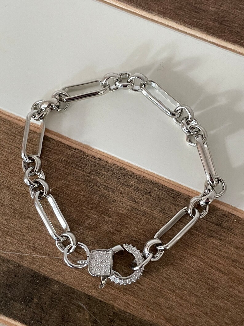 May include: Silver-toned chain bracelet with a rectangular link design. The bracelet features a decorative clasp with a square center and a surrounding ring, both encrusted with small, clear stones. The bracelet is displayed on a white and brown surface.