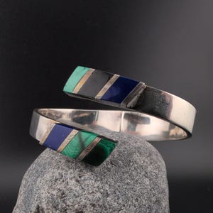 Op de afbeelding: Een bypass-stijl armband gemaakt van gepolijst zilverkleurig metaal. De twee uiteinden zijn voorzien van ingelegde diagonale segmenten van groene malachiet, zwarte onyx en diepblauwe lapis lazuli, wat een geometrisch patroon vormt.