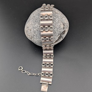 Könnte beinhalten: Ein silberfarbenes Armband mit geometrischem Design, das auf einem grauen Stein liegt. Das Armband hat rechteckige und sechseckige Glieder, die durch kleine Kettenglieder verbunden sind. Der Verschluss ist sichtbar, und das Armband scheint Vintage zu sein.