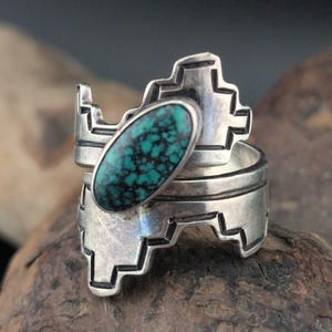 May include: A sterling silver ring featuring a central oval turquoise stone with dark matrix patterns. The band has a wide, stepped geometric design with oxidised recessed lines that create a sharp contrast against the polished silver surface.