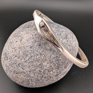 May include: A silver-toned bracelet with a small, round, clear gemstone. The bracelet has an open design with a curved shape and a decorative element. The bracelet is resting on a gray stone against a black background.