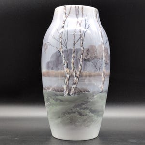Könnte beinhalten: Eine weiße Keramikvase mit einer handgemalten Landschaftsszene. Das Design zeigt drei schlanke Birken im Vordergrund sowie ein graubraunes Flussufer, entfernte Bäume und einen weichen Himmel in verschiedenen Grau- und Grüntönen auf dem Gefäß.