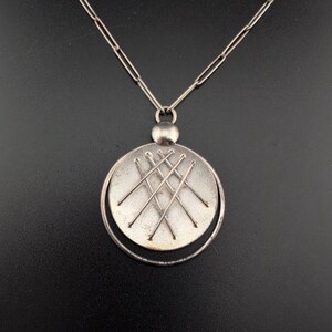 Könnte beinhalten: Eine silberfarbene Anhängerkette. Der Anhänger hat ein kreisförmiges Design mit strukturierter Oberfläche und sich kreuzenden Linien. Die Halskette hat eine zarte Kette und einen kleinen, runden Verschluss. Der Schmuck wird vor dunklem Hintergrund präsentiert.