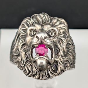 Peut inclure: Bague tête de lion argentée avec une pierre précieuse rouge rubis dans la gueule. La bague présente des textures détaillées dans la crinière et le visage du lion, créant un design audacieux. Le rubis ajoute une touche de couleur.