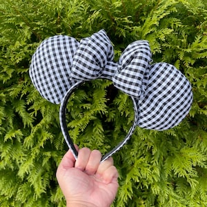 May include: Black and white gingham patterned mouse ears headband. The ears are round and the bow is large and centered. The headband is black with white trim. The background is green foliage.