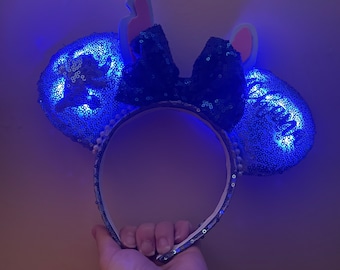 Könnte beinhalten: Blaue leuchtende Minnie Mouse Ohren mit einer schwarzen Schleife und blauen und weißen Paillettendetails. Die Ohren sind in Form von Mickey Mouse Ohren und haben eine hellblaue Schleife oben.