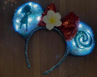 May include: Light-up mouse ears with blue sequined ears, a red sequined bow, and a white flower. One ear features a silhouette of a character, the other a spiral design. The headband is teal.