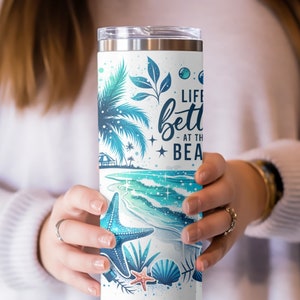 May include: A white stainless steel tumbler with a blue and green beach scene and the text "Life is better at the beach".