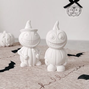 Könnte beinhalten: Zwei weiße Keramik-Kürbisfiguren mit bemalten Gesichtern. Der Kürbis links hat einen Hexenhut und der Kürbis rechts ein breites Grinsen. Die Kürbisse stehen auf einer braunen Jutestoff-Oberfläche mit einem schwarzen Fledermaus-Design.