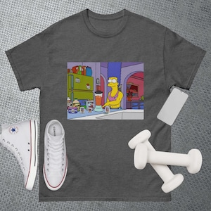 May include: A gray t-shirt with a cartoon image of a yellow-haired woman in a kitchen making a smoothie. The woman is wearing a blue tank top and has a muscular physique. The image is surrounded by a white border.