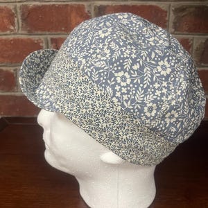 May include: A cap featuring a blue and white floral pattern on the crown and a contrasting band with a smaller, dense blue and white floral print. The cap has a short, curved brim and is displayed on a white mannequin head form.