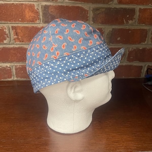 Puede incluir: Una gorra de tela azul con un patrón de cachemira rojo en la corona y una banda azul en contraste con un estampado floral blanco. La gorra tiene una visera corta hacia arriba y costuras decorativas en zigzag de color blanco.