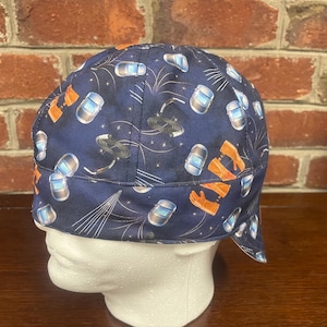 May include: A navy blue welding cap featuring a repeating pattern of silver welding helmets, orange protective gloves, and white line graphics representing sparks or light rays. The cap has a rounded crown and a short, stiffened brim at the back.