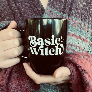 May include: Black ceramic mug with the words "Basic Witch" in white script. The mug has a curved handle and is held by a person wearing a burgundy and gray sweater.