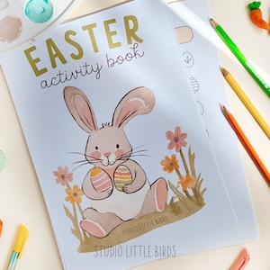 May include: An Easter activity book with a bunny illustration holding decorated eggs. The cover reads "EASTER activity book" in gold lettering. Colored pencils, markers, and decorative carrots and eggs are scattered around the book.