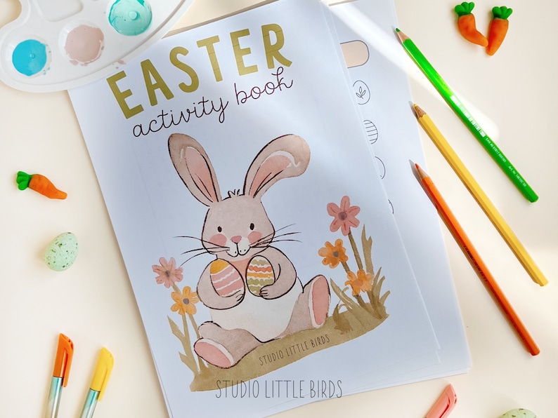 May include: An Easter activity book with a bunny illustration holding decorated eggs. The cover reads "EASTER activity book" in gold lettering. Colored pencils, markers, and decorative carrots and eggs are scattered around the book.