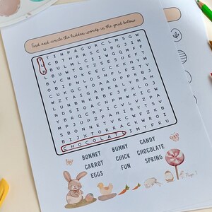 May include: Easter-themed word search puzzle on white paper with a bunny illustration, a carrot, and a lollipop. The word "chocolate" is circled. Colored pencils and paint palette are in the background.