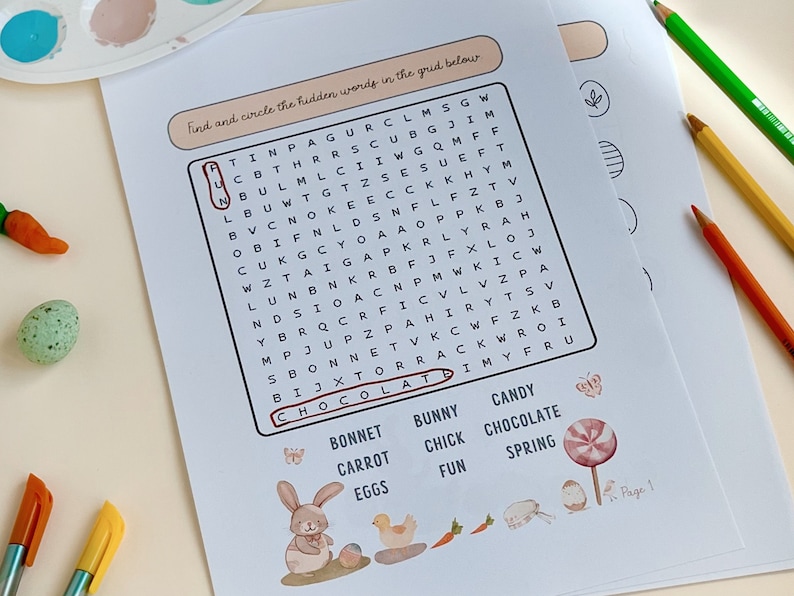 May include: Easter-themed word search puzzle on white paper with a bunny illustration, a carrot, and a lollipop. The word "chocolate" is circled. Colored pencils and paint palette are in the background.
