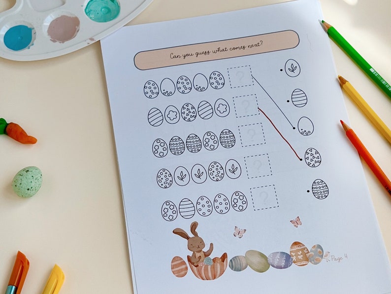 May include: Easter-themed activity sheet with egg patterns and the text "Can you guess what comes next?" A bunny illustration and colorful eggs are at the bottom. Colored pencils and paint palette are in the background.