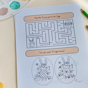 May include: A white paper with a maze and the text "Help the Bunny get to the Eggs". Below the maze is the text "Can you spot 10 differences?". The page also features illustrations of bunnies and Easter eggs. Colored pencils and paint are in the background.