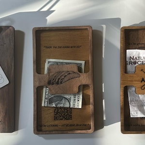 Puede incluir: Tres organizadores de madera con acabado oscuro. El primero es un portapapeles con clip metálico y tarjeta. El segundo tiene ranuras para efectivo y un objeto pequeño. El tercero tiene ranuras para recibos y un logotipo.