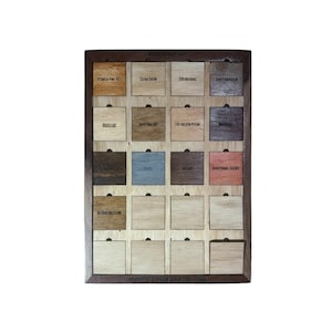 May include: A wooden display board featuring various wood stain test samples. Each sample is a small, square block with a different color and finish, labeled with names like "Powder Pine 421" and "Traditional Cherry". The board is framed in dark wood.