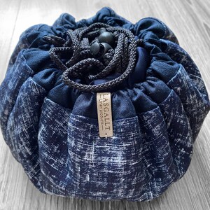 May include: A navy blue drawstring makeup bag with a white textured pattern. The bag is round and gathered at the top with a black drawstring and toggle. A small tag reads "LASGALLT Linen Products".