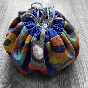 May include: A colorful drawstring bag with a blue lining and a label that says "SCALIT". The bag is made of a fabric with a geometric pattern in shades of yellow, orange, green, blue, and purple.