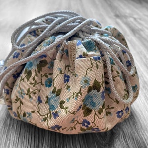 May include: A white drawstring bag with a floral pattern featuring blue flowers and green leaves. The bag has a light grey drawstring and a brown label that reads "ASGALLI - Linen Products".