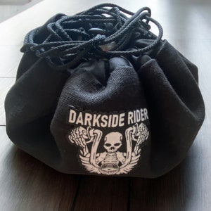 以下が含まれることがあります： 白いドクロと交差した骨のグラフィックと「DARKSIDE RIDER」のテキストが書かれた黒い巾着袋。