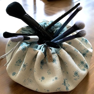 Op de afbeelding: Een witte en blauwe bloemen drawstring make-up tas met een zwarte trekkoord en verschillende make-up kwasten erin.