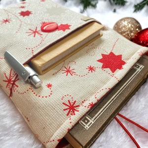 May include: A cream-colored book sleeve with a red star and snowflake pattern, holding two books. A silver pen is tucked into the sleeve. A red button and red ribbon add a festive touch. Christmas ornaments are in the background.