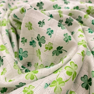 May include: White fabric featuring a repeating pattern of green shamrocks. The shamrocks are in various shades of green and are spread across the fabric. The fabric has a textured appearance, possibly linen or a similar weave.