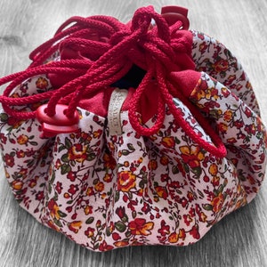 May include: A red drawstring bag with a floral print. The bag is made of fabric and has a red drawstring closure. The bag is sitting on a wooden surface.
