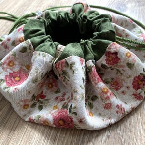 May include: A round drawstring bag with a floral pattern in pink and red on a cream background. The top and drawstring are green. The bag is made of fabric and sits on a wooden surface.
