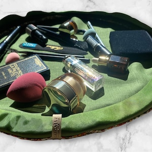 May include: A green makeup bag with a floral pattern and the text "LASSIGALLE" on the bottom. The bag is filled with various makeup products, including brushes, sponges, and containers of cream and liquid.