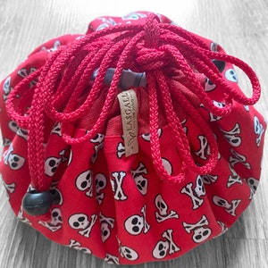Pode incluir: Saco de cordão vermelho com um padrão de caveira e ossos cruzados. O saco é feito de tecido e tem um fecho de cordão. O saco é amarrado com um cordão vermelho e tem um botão preto.