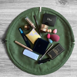May include: A green round makeup organizer with various makeup items, including a black eyeshadow palette labeled "Dior", a clear bottle with a gold cap, a black eyeliner pencil, a silver nail file, a white makeup sponge, a black and gold makeup brush, a black and gold mascara, a black and gold eyeliner, a black and gold makeup palette labeled "L'OREAL PARIS COLOR RICHE LA PALETTE MATTE", and a black and gold makeup palette labeled "Dior".
