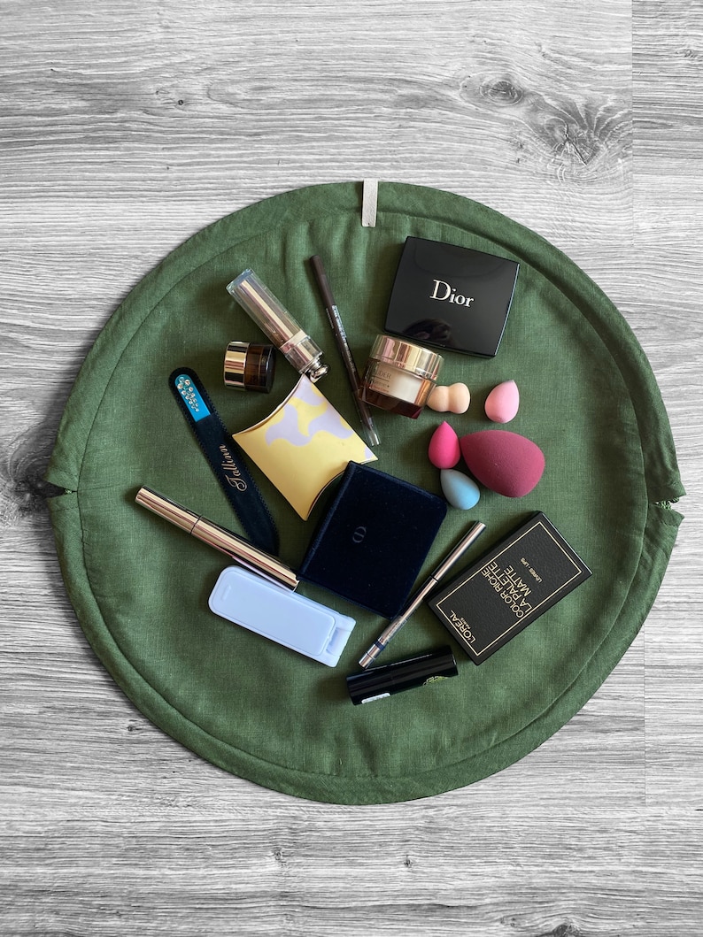 May include: A green round makeup organizer with various makeup items, including a black eyeshadow palette labeled "Dior", a clear bottle with a gold cap, a black eyeliner pencil, a silver nail file, a white makeup sponge, a black and gold makeup brush, a black and gold mascara, a black and gold eyeliner, a black and gold makeup palette labeled "L'OREAL PARIS COLOR RICHE LA PALETTE MATTE", and a black and gold makeup palette labeled "Dior".